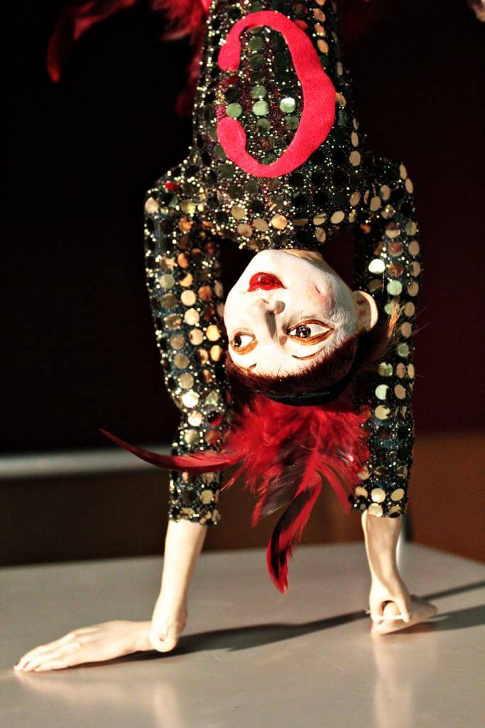 Zdjecie do spektaklu Abecadło. Na zdjęciu lalka tancerka, ubrana w strój z cekinów i pióra na głowie. Lalka stoi na rękach.