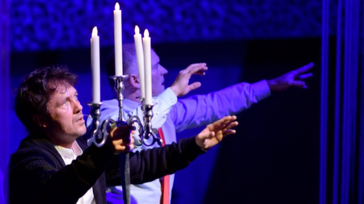 Zdjęcie do spektaklu Dobroć nasza dobroć. Dwóch mężczyzn z wyciagnietymi przed siebie rękoma. jeden trzyma świecznik z piecioma świecami.
