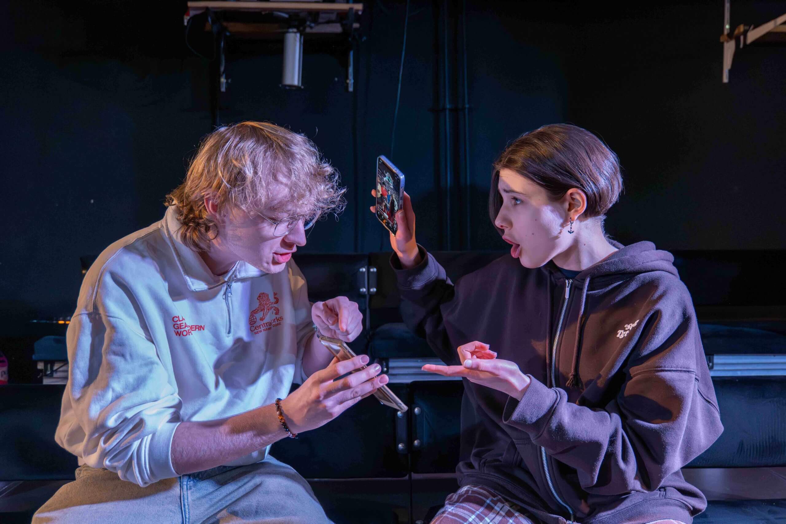 Na zdjęciu dwoje aktorów, po lewej chłopak w białej bluzie, po prawo dziewczyna w szarej bluzie. gestykulują jak by się o coś kłócili.