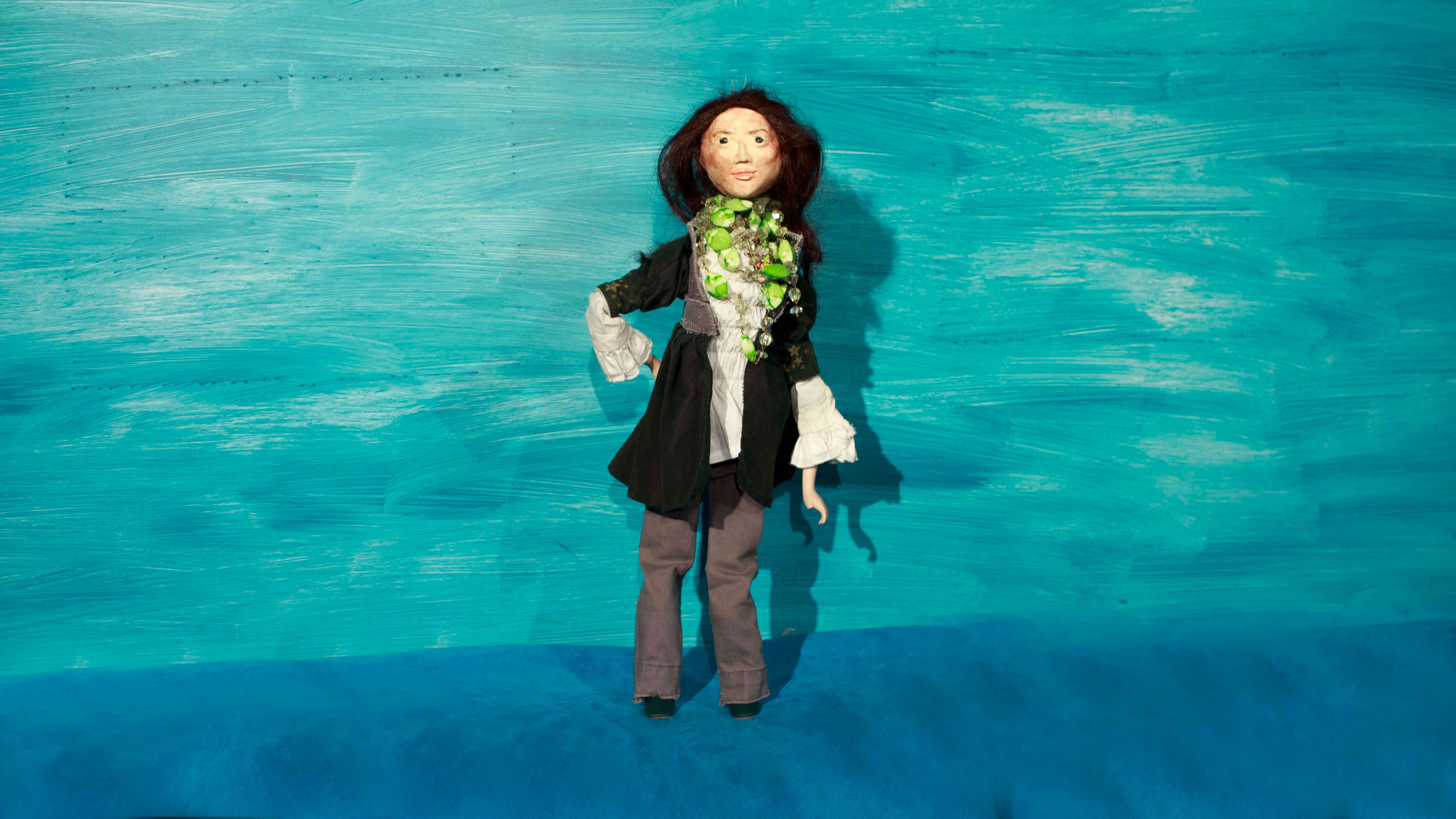 zdjęcie przedstawiające stojącą lalkę przedstawiającą kobietę w ciemnych długich włosach, o ciemnych oczas, ubraną w białą koszulę, ciemny sweter, szare spodnie i czarne buty, z zielonymi i przezroczystymi koralami wokół szyi, prawą ręką podpiera się pod bok, niebieskie tło