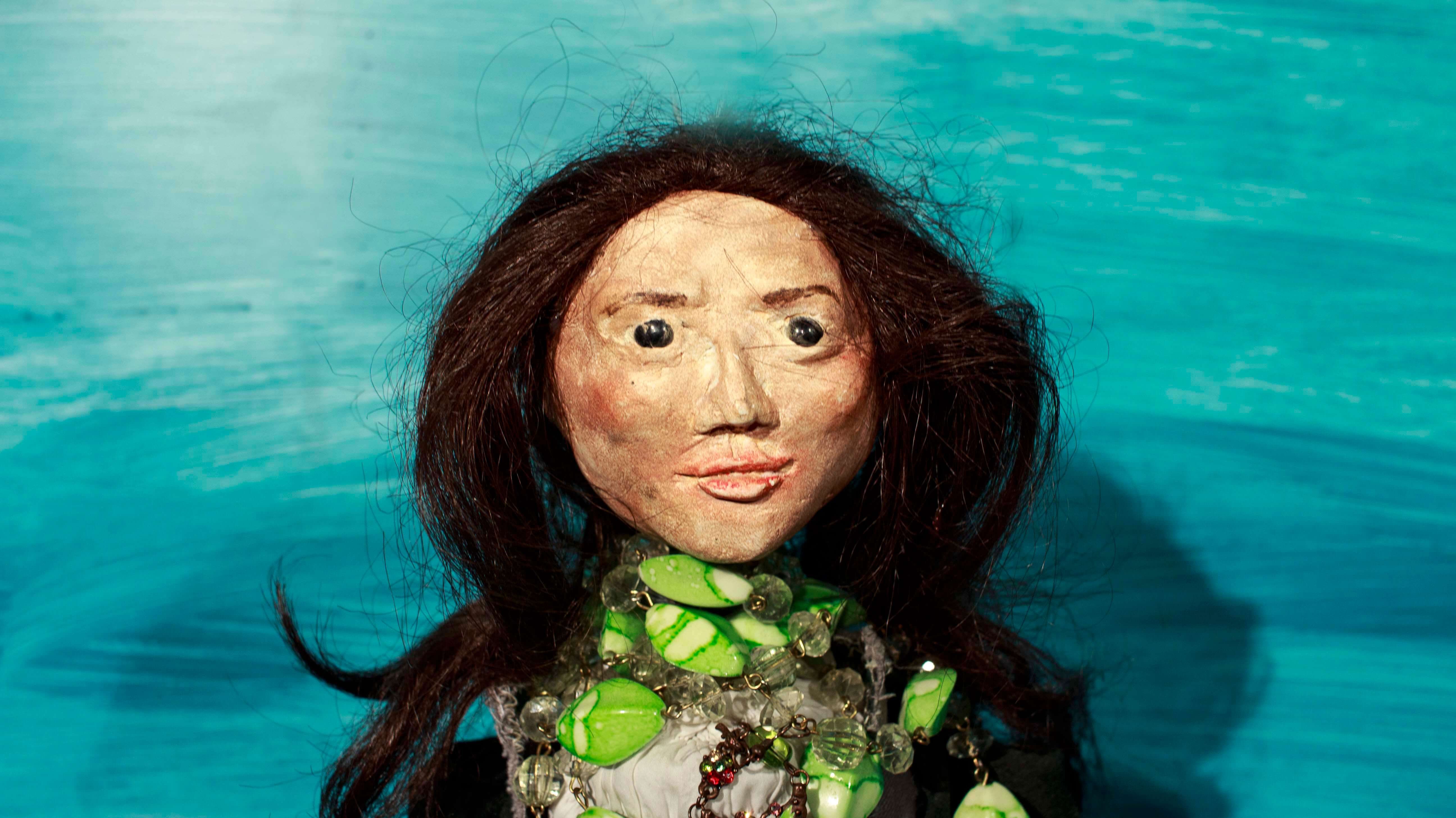 zbliżenie na twarz lalki przedstawiającej kobietę o ciemnych rozpuszczonych włosach, czarnych oczach, z zielonymi i przezroczystymi koralami wokół szyi, niebieskie tło