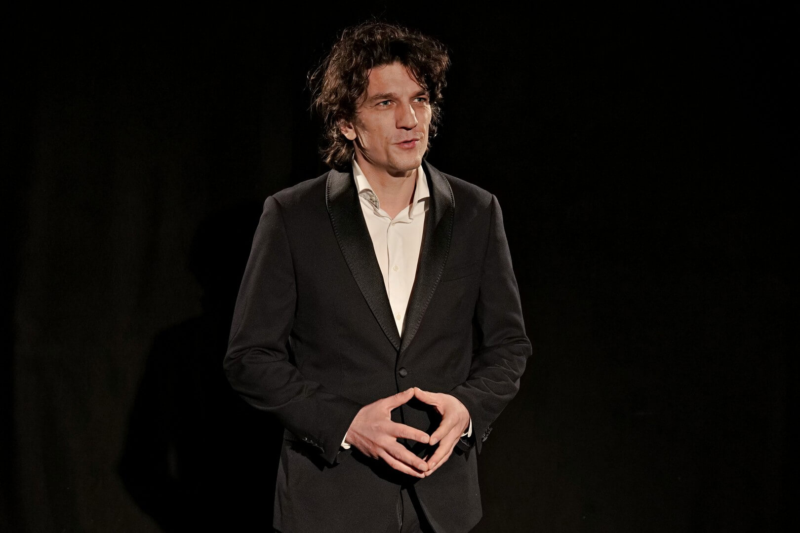 na zdjęciu mężczyzna w czarnym garniturze i białej koszuli, widziany od pasa w górę, dłonie złączone na wysokości pasa