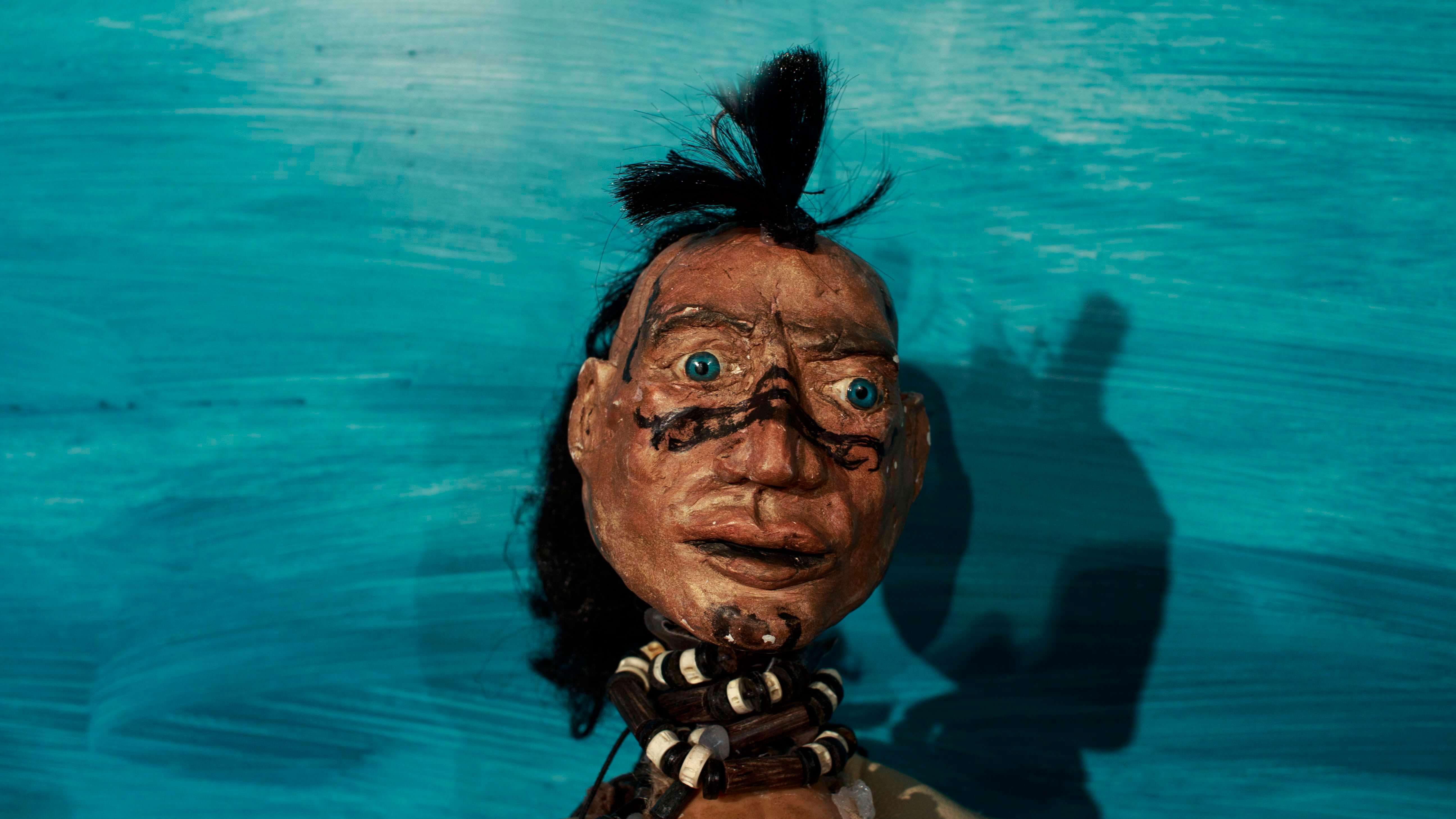 głowa lalki o ciemnym kolorze skóry, czarnych włosach, i niebieskich oczach, na nosie i policzkach ma narysowane czarne znaki, włosy częścio związane na czubku głowy, na szyi korale, niebieskie tło