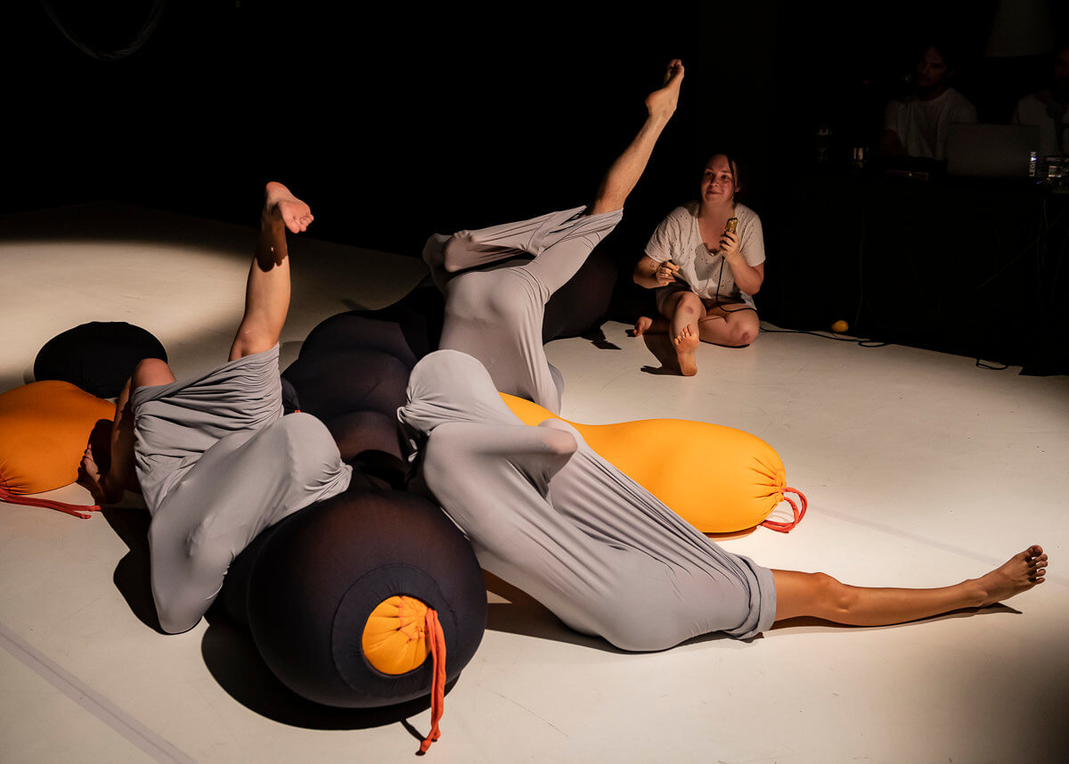 Zdjęcie przedstawia grupę tancerzy, leżących na podłodze owinietych w szary materiał tak że widoczne są tylko nogi.