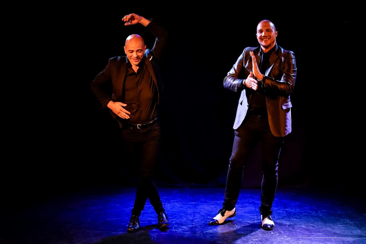 dwóch mężczyzn stoi na scenie, ubrani w czarne spodnie, czarne marynarki, czarne koszule i eleganckie buty, mężczyzna po lewej tańczy ma ugięte w kolanach nogi, jedną ręka trzyma się pod bok, drugą unosi nad głowę, drugi mężczyzna stoi, trzyma przed sobą złączone dłonie, uśmiecha się, tło jest ciemne, świeci niebieskie światło, zdjęcie z wydarzenia w teatrze gdynia główna