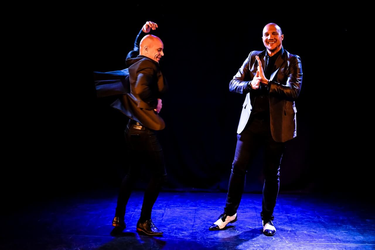 dwóch mężczyzn stoi na scenie, ubrani w czarne spodnie, czarne marynarki, czarne koszule i eleganckie buty, mężczyzna po lewej tańczy ma ugięte w kolanach nogi, jedną ręka trzyma się pod bok, drugą unosi nad głowę, drugi mężczyzna stoi, trzyma przed sobą złączone dłonie, uśmiecha się, tło jest ciemne, świeci niebieskie światło, zdjęcie z wydarzenia w teatrze gdynia główna