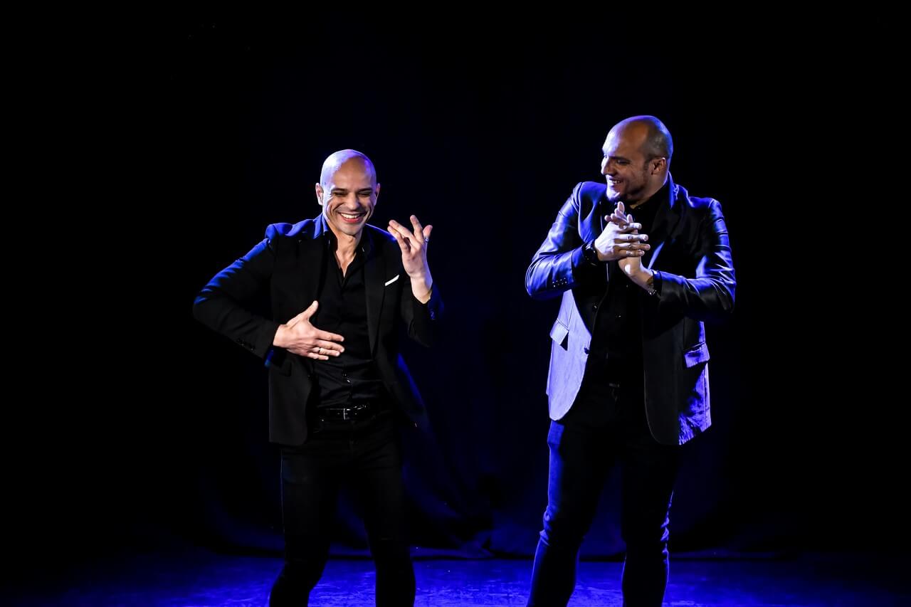 dwóch mężczyzn na scenie, po lewej mężczyzna tańczy, ma ugięte nogi w kolanach, ręce zgięte w łokciach , ma radosny wyraz twarzy, ubrany w czarne spodnie, czarną koszulę, czarną marynarkę, jest łysy, po prawej stoi łysy mężczyzna z lekkim zarostem, patrzy w obiektyw, ma złożone przed sobą dłonie, ubrany podobnie jak pierwszy mężczyzna, uśmiecha się, tło jest ciemne, świeci niebieskie światło, zdjęcie z wydarzenia w teatrze gdynia główna