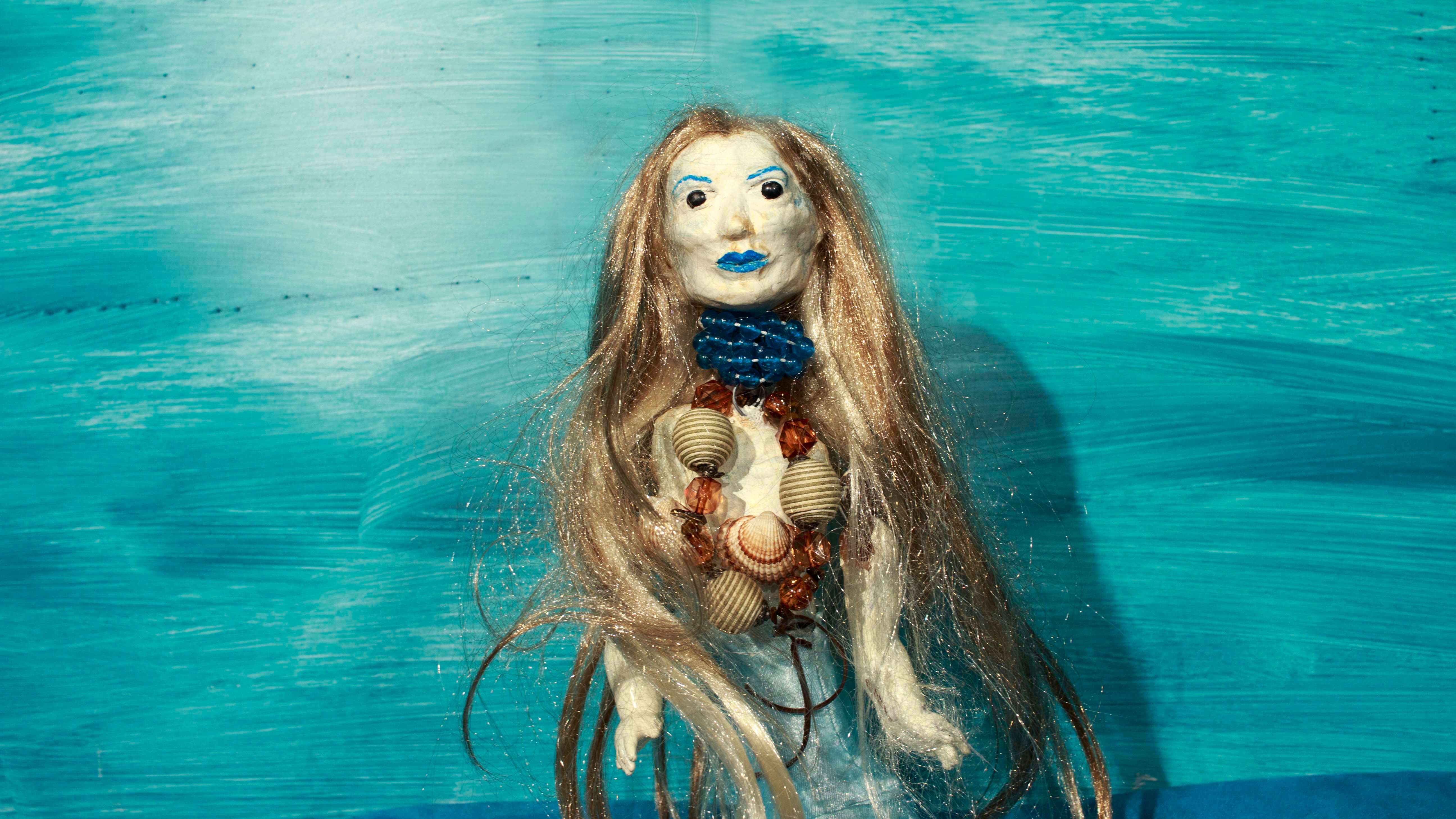 lalka postaci syreny ma długie splątane włosy, jasną cerę, niebieskie usta i brwi, na szyi niebieskie korale oraz naszyjnik z muszli i bursztynu, czarne oczy, niebieskie tło