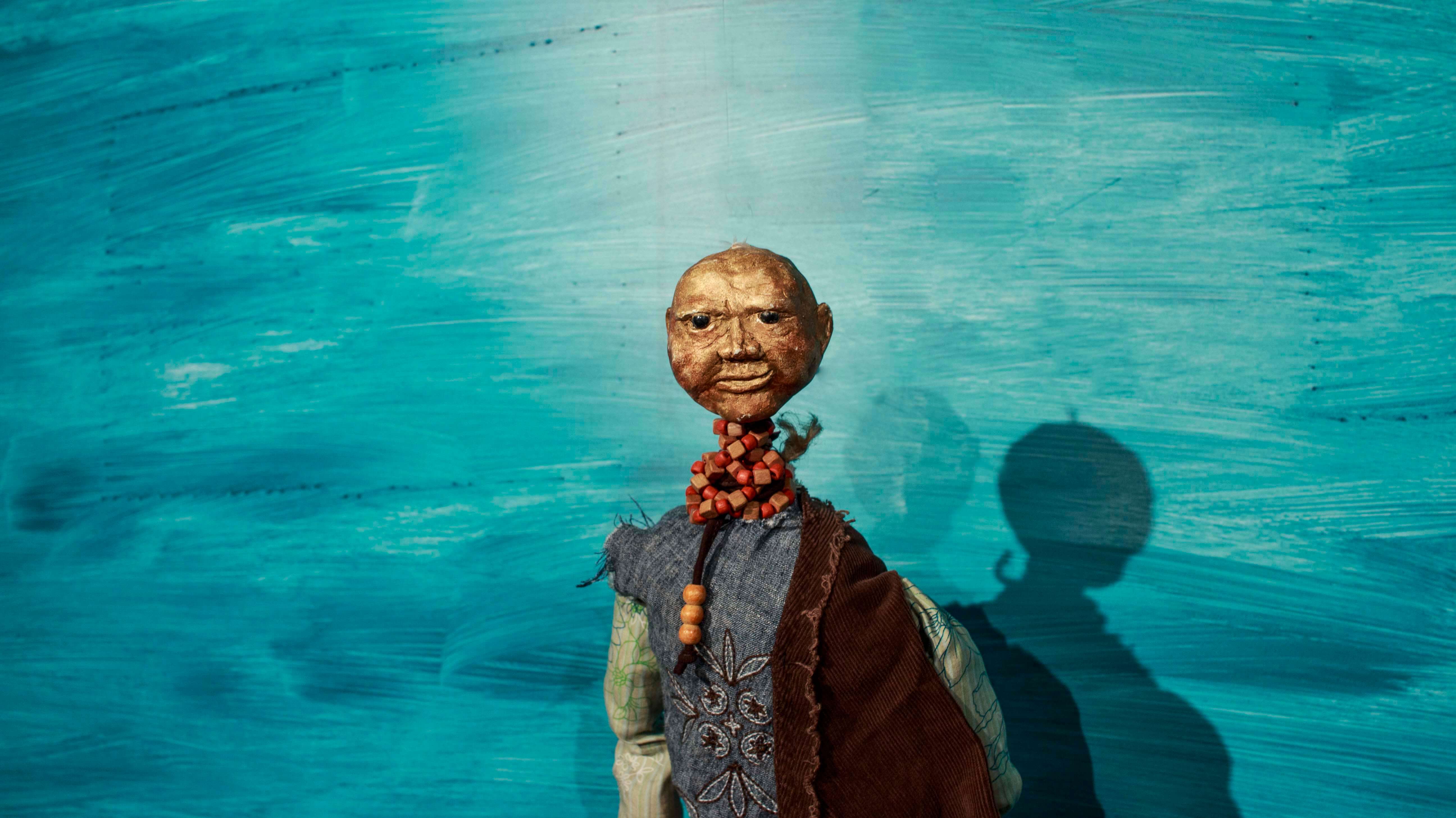 lalka, łysa, starsza postać ubrana w niebiesko szarą koszulę i brązowy przerzucony przez ramię materiał, na szyi ma korale, niebieskie tło
